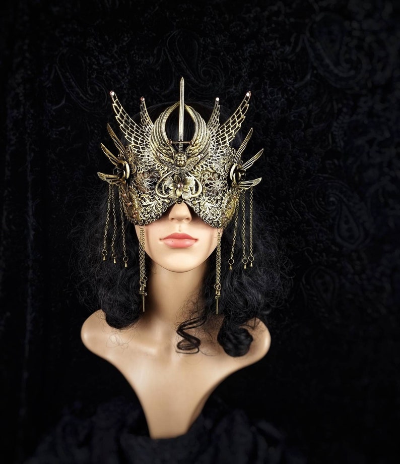 May include: A gold masquerade mask with intricate details and dangling chains. The mask is worn by a mannequin with black hair.