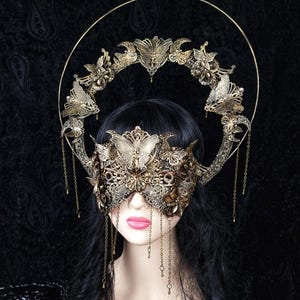 May include: An ornate gold-toned filigree mask featuring butterfly and floral motifs, paired with a matching halo-style headpiece. The headpiece includes a large circular wire frame adorned with butterflies and delicate hanging metal chains.