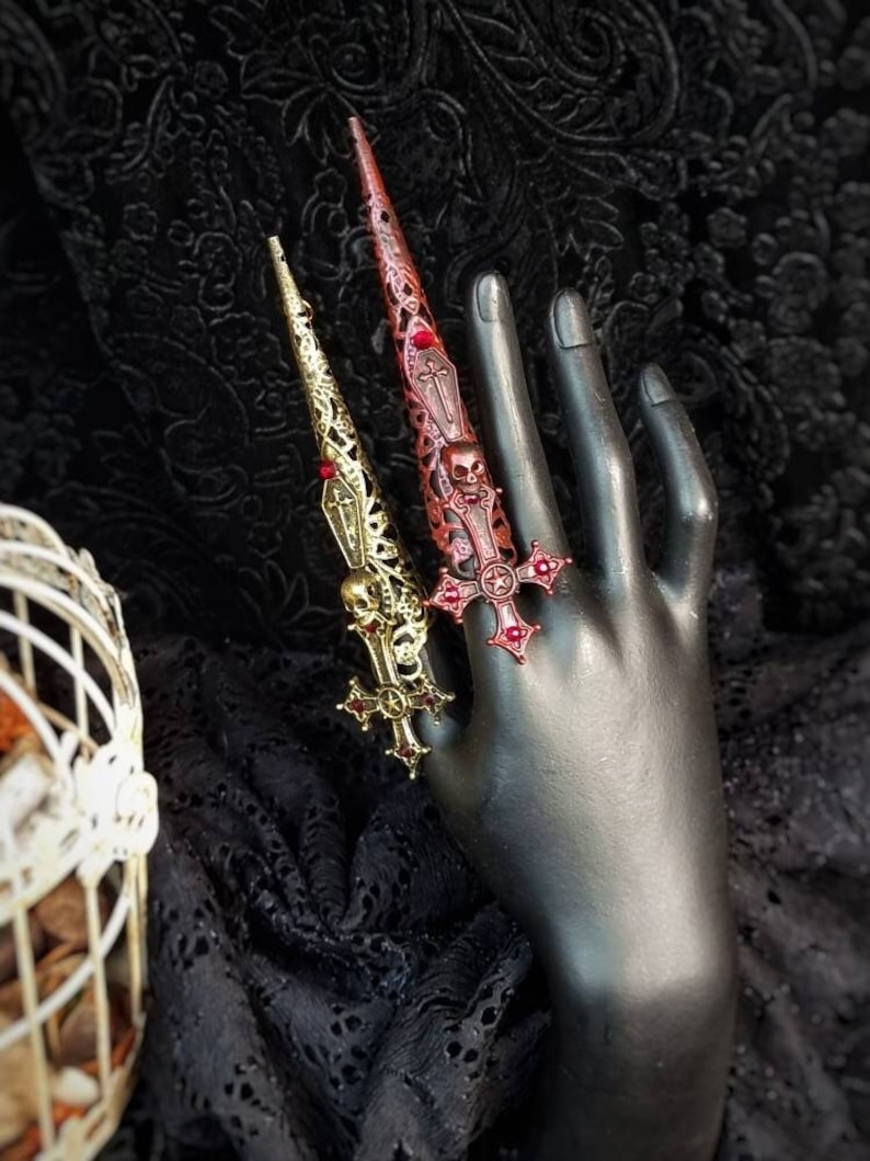 May include: Two long, pointed finger armor rings featuring intricate filigree metalwork. One ring is gold-toned with a skull and cross motif, while the other is dark red with a skull and cross design. Both pieces are adorned with small red accents.