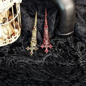 May include: Two long, pointed finger armor rings featuring intricate metalwork. One ring is antique gold-toned, and the other is dark red-toned. Both designs include a skull, a coffin, and a cross motif with small red embedded stones.