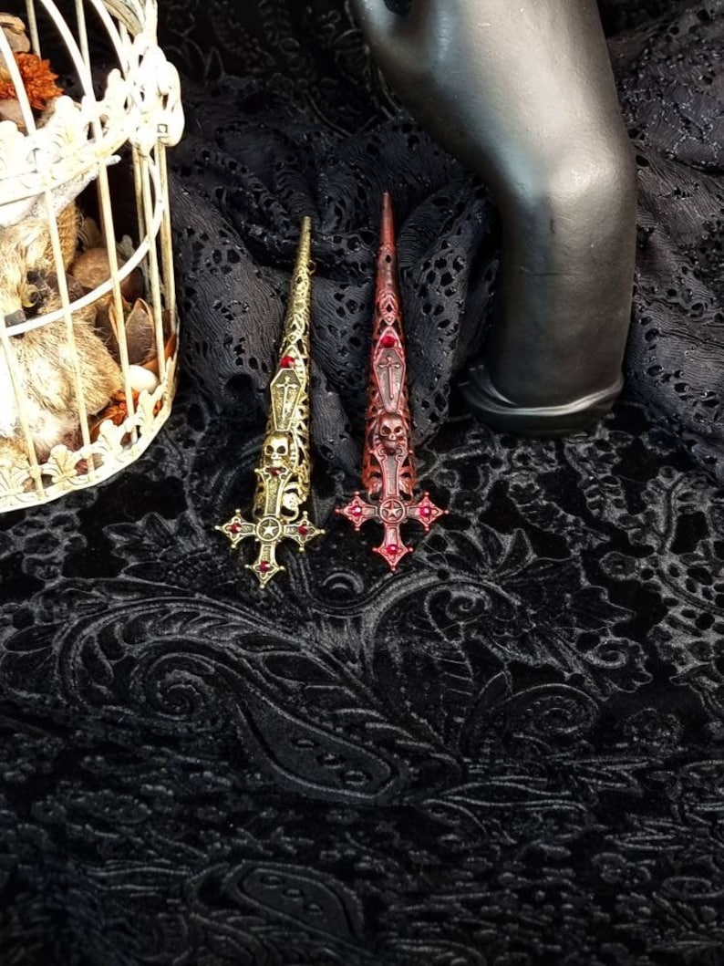 May include: Two long, pointed finger armor rings featuring intricate metalwork. One ring is antique gold-toned, and the other is dark red-toned. Both designs include a skull, a coffin, and a cross motif with small red embedded stones.