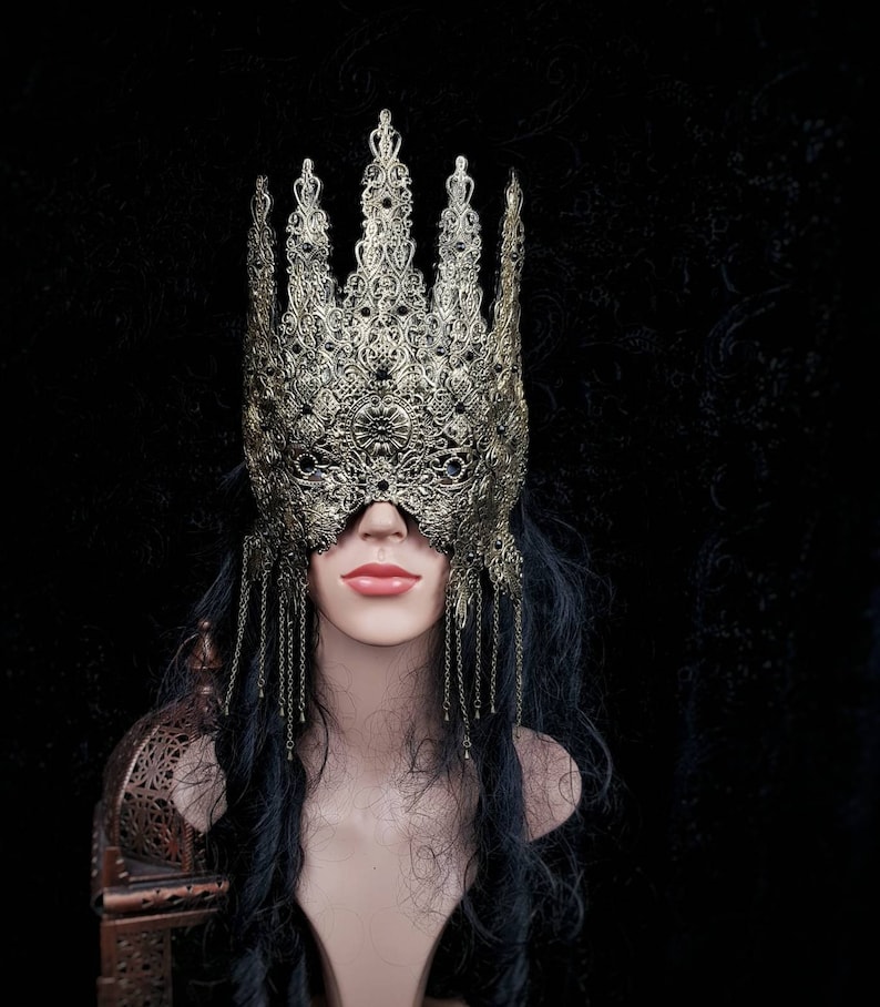 Puede incluir: Una m&aacute;scara de metal dorado para un baile de m&aacute;scaras con detalles intrincados y cadenas colgantes. La m&aacute;scara cubre los ojos y la frente y tiene una corona puntiaguda.