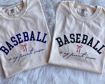 May include: Two cream colored t-shirts with the text "Baseball is my favorite season" in blue and black lettering. A red bow is embroidered above the text on each shirt.