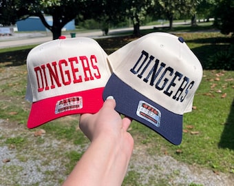 May include: Two baseball caps with the word "DINGERS" embroidered on the front. One cap has a red brim and lettering, the other has a navy blue brim and lettering. Both caps have a cream-colored crown.