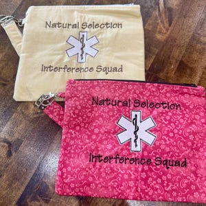 May include: Two zippered pouches, one cream-colored and one pink, are displayed. Both pouches feature the text "Natural Selection" and "Interference Squad" with a white embroidered medical symbol. The pink pouch has a floral pattern.