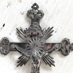 May include: An ornate silver crucifix depicting Jesus Christ. The cross has detailed designs, including a sunburst and the letters INRI. The background is a distressed white surface.