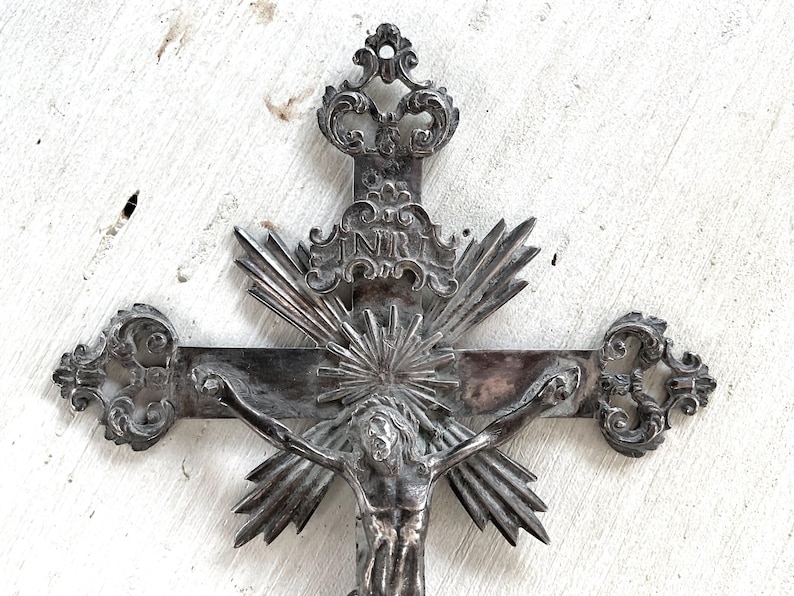 May include: An ornate silver crucifix depicting Jesus Christ. The cross has detailed designs, including a sunburst and the letters INRI. The background is a distressed white surface.
