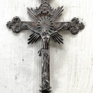 May include: An ornate silver crucifix depicting Jesus Christ. The cross has decorative elements, including a sunburst design behind the figure and a skull and crossbones at the base. The design is intricate and detailed, with a vintage aesthetic.