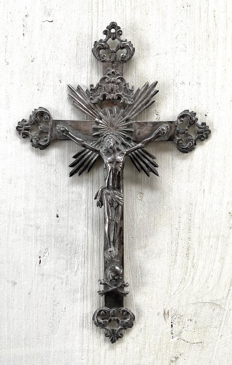 May include: An ornate silver crucifix depicting Jesus Christ. The cross has decorative elements, including a sunburst design behind the figure and a skull and crossbones at the base. The design is intricate and detailed, with a vintage aesthetic.