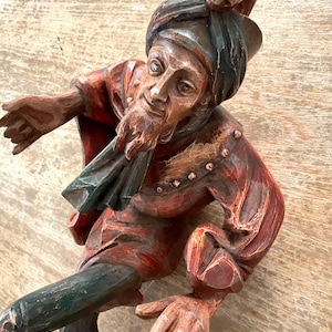 Puede incluir: Una figura de madera tallada de un hombre con turbante y abrigo rojo con detalles verdes. La figura tiene una larga barba y una pequeña cara tallada en el turbante. La figura está posada en una posición dinámica e inclinada.