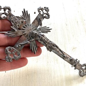 May include: An antique silver crucifix with intricate detailing. The cross features a detailed depiction of Jesus Christ and decorative flourishes at the ends of the cross arms. The design includes a sunburst pattern behind the figure.