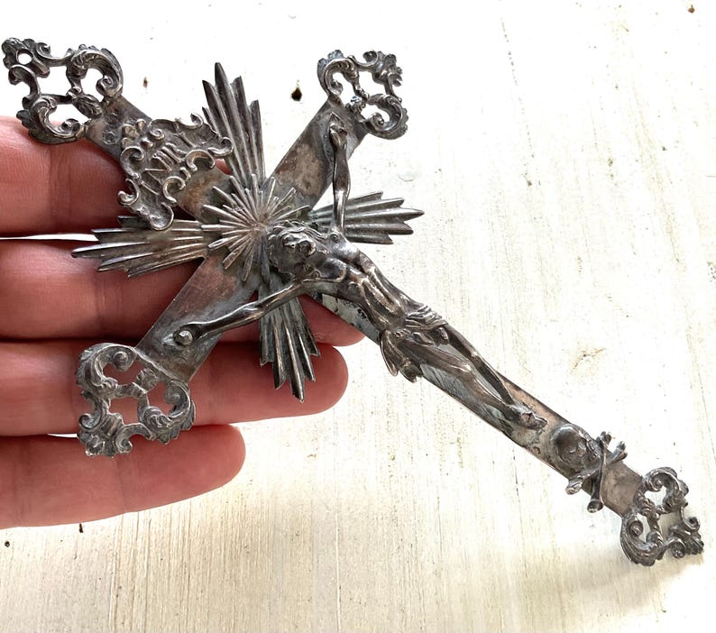 May include: An antique silver crucifix with intricate detailing. The cross features a detailed depiction of Jesus Christ and decorative flourishes at the ends of the cross arms. The design includes a sunburst pattern behind the figure.