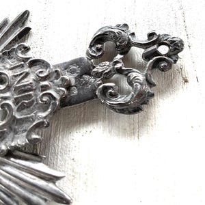 May include: Close-up of a silver-toned metal cross with ornate, swirling details. The cross features the letters "INRI" and a decorative, stylized design. The cross is set against a white, textured background.