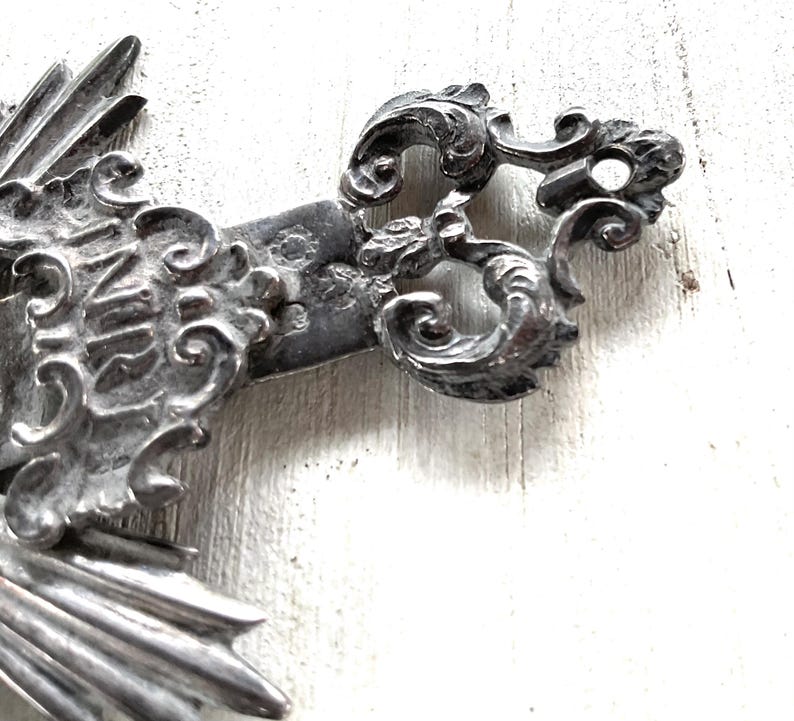 May include: Close-up of a silver-toned metal cross with ornate, swirling details. The cross features the letters "INRI" and a decorative, stylized design. The cross is set against a white, textured background.