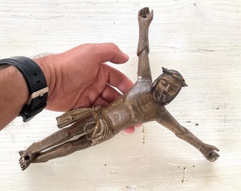 May include: A bronze-toned sculpture of Jesus Christ on the cross, with arms outstretched. The figure wears a crown of thorns and a loincloth. The sculpture is held by a hand.