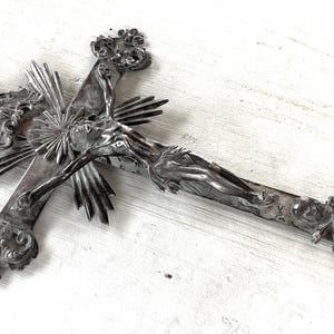 May include: An ornate silver crucifix with detailed carvings. The cross features a central figure and sunburst design, with decorative flourishes at the ends. A religious item with an antique aesthetic.
