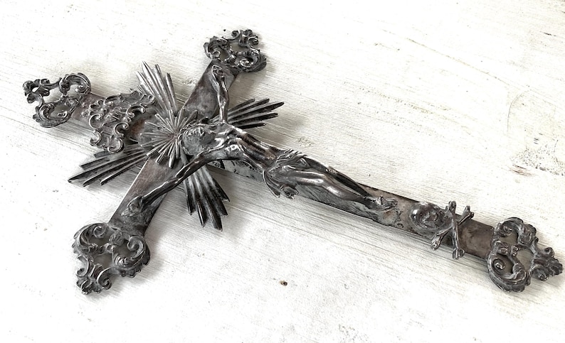 May include: An ornate silver crucifix with detailed carvings. The cross features a central figure and sunburst design, with decorative flourishes at the ends. A religious item with an antique aesthetic.