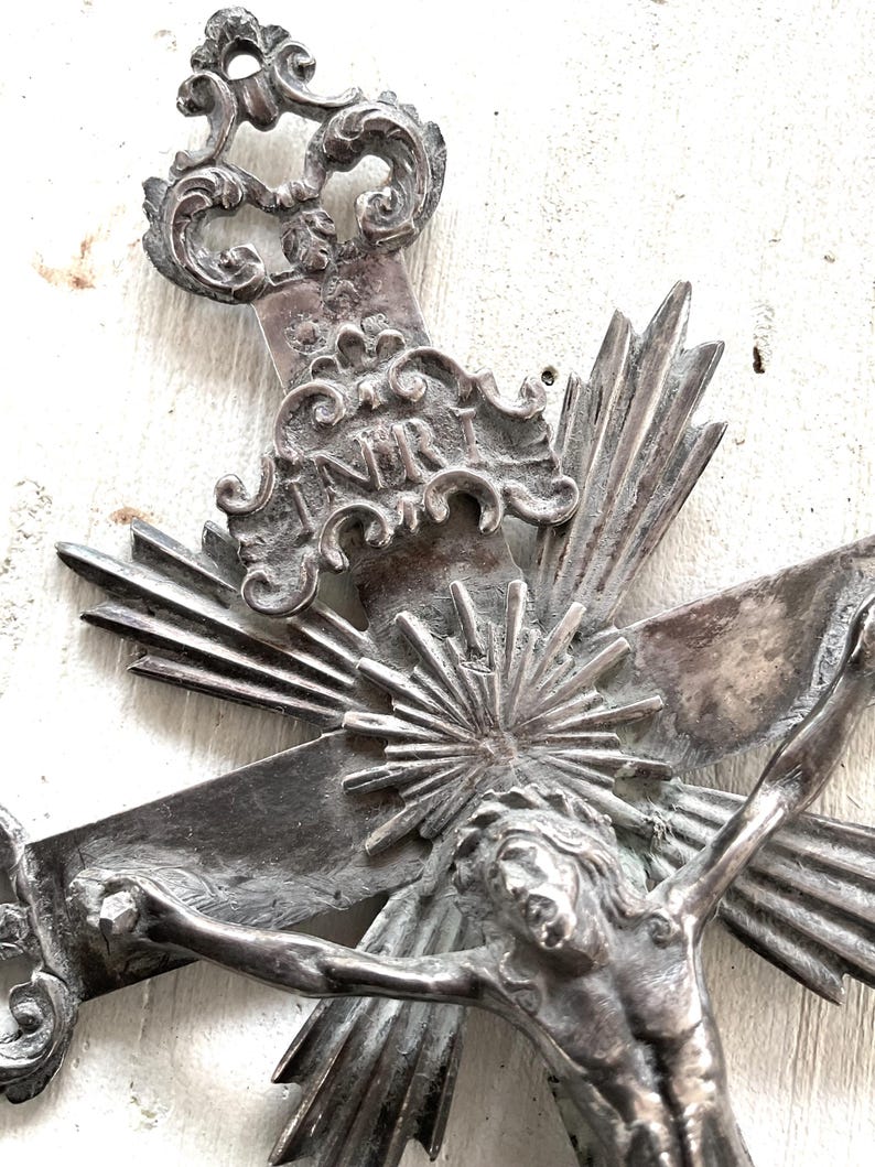 May include: An antique silver-coloured crucifix with intricate detailing. The cross depicts Jesus Christ, with the inscription "INRI" above. The design includes ornate embellishments and sunburst rays emanating from behind the figure.