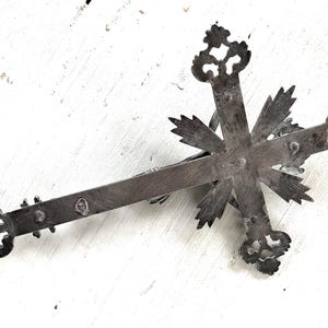 May include: An antique silver-coloured crucifix with intricate, stylized floral patterns at the ends of the cross arms. The cross features a central figure and is set against a white, textured background.