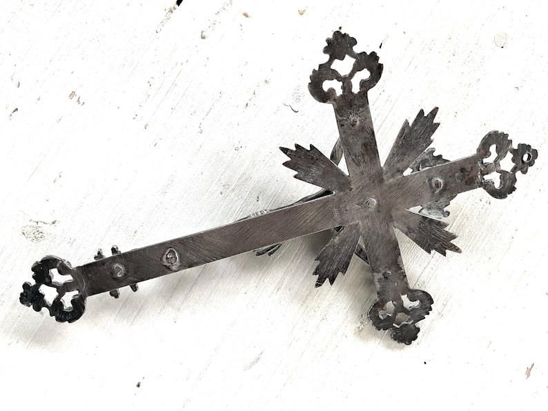 May include: An antique silver-coloured crucifix with intricate, stylized floral patterns at the ends of the cross arms. The cross features a central figure and is set against a white, textured background.