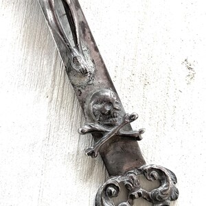 May include: An antique silver-coloured dagger with religious and gothic details. The blade features a sculpted figure, a skull with crossed bones, and ornate scrollwork. The dagger is set against a white background.