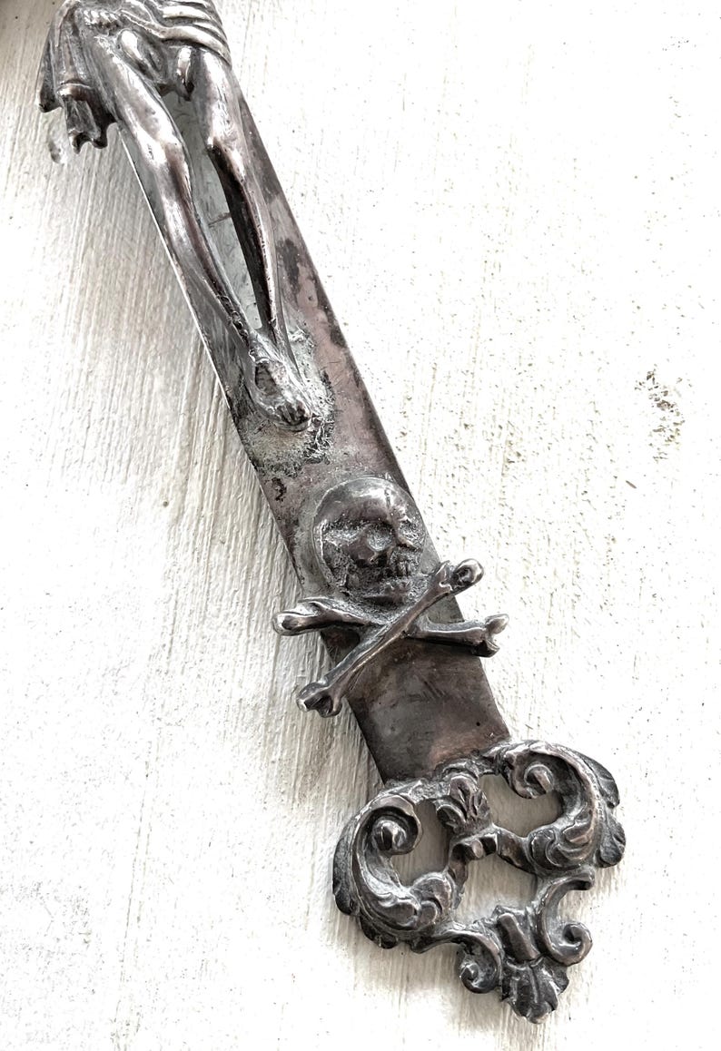 May include: An antique silver-coloured dagger with religious and gothic details. The blade features a sculpted figure, a skull with crossed bones, and ornate scrollwork. The dagger is set against a white background.