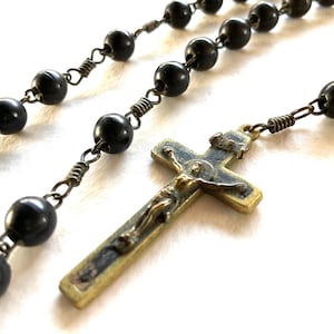 Puede incluir: Primer plano de un rosario con cuentas negras y un crucifijo de bronce. Las cuentas están unidas con alambre, y el crucifijo presenta una representación detallada de Jesucristo. El rosario está sobre un fondo blanco.