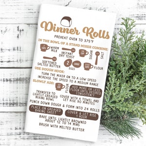 May include: A white kitchen towel with brown text and illustrations showing the ingredients and steps for making dinner rolls. The text includes "Dinner Rolls", "Preheat oven to 375&deg;F", "In the bowl of a stand mixer combine:", "Warm milk", "Instant dry yeast", "Softened salted butter", "2 large eggs", "5 1/2 cups flour", "Sugar", "Salt", "Use dough hook:", "Turn the mixer on to a low speed", "Increase the speed to a medium range", "Slowly add:", "Flour", "Transfer to lightly greased mixing bowl", "Add more or less flour until desired consistency", "Cover with a towel and let rise 90 minutes", "Punch down dough & form into 24 rolls", "Place on greased baking sheet", "Cover & let rise 1 hour", "Bake until lightly browned about 12 to 14 mins", "Brush with melted butter".