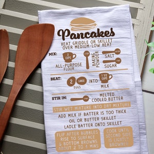 May include: White kitchen towel with brown text and illustrations showing how to make pancakes. The text includes "Pancakes", "Heat griddle or skillet over medium-low heat", "Mix", "Beat", "Stir in", "Stir wet mixture into dry mixture", "Add milk if batter is too thick", "Oil or butter skillet", "Ladle batter onto skillet", "Flip after bubbles rise to surface & bottom browns about 2 to 4 mins", "Cook until second side is lightly browned", "2 cups all-purpose flour", "2 large eggs", "1 1/2 cups milk", "Melted cooled butter", "Salt", "Sugar", "Baking powder".