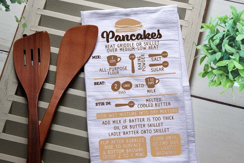May include: White kitchen towel with brown text and illustrations showing how to make pancakes. The text includes "Pancakes", "Heat griddle or skillet over medium-low heat", "Mix", "Beat", "Stir in", "Stir wet mixture into dry mixture", "Add milk if batter is too thick", "Oil or butter skillet", "Ladle batter onto skillet", "Flip after bubbles rise to surface & bottom browns about 2 to 4 mins", "Cook until second side is lightly browned", "2 cups all-purpose flour", "2 large eggs", "1 1/2 cups milk", "Melted cooled butter", "Salt", "Sugar", "Baking powder".