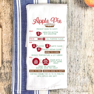 May include: White tea towel with a red and green apple pie recipe printed on it. The recipe includes instructions for melting, stirring, adding, filling, and baking the pie. The text also includes measurements in cups and ounces.