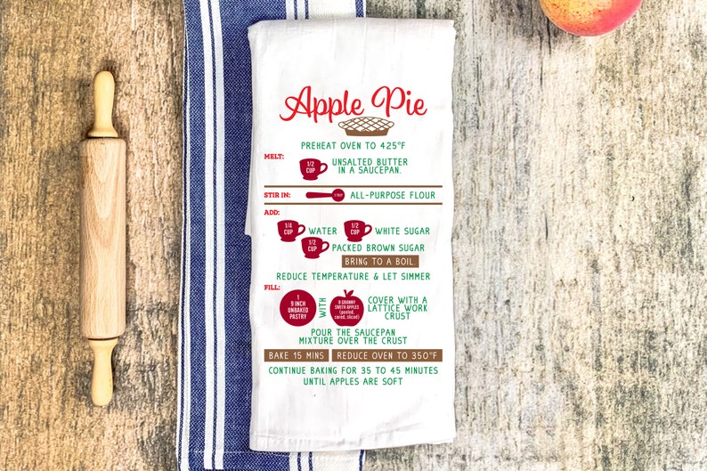 May include: White tea towel with a red and green apple pie recipe printed on it. The recipe includes instructions for melting, stirring, adding, filling, and baking the pie. The text also includes measurements in cups and ounces.