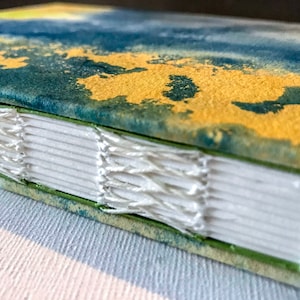 Peut inclure: Gros plan sur le dos d'un livre fait main avec une couverture verte. Le livre est relié avec du fil blanc et a une couverture en papier marbré bleu et or.