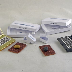 May include: Miniature gold and black laptops with open screens displaying colorful images, alongside white boxes labeled "MacBook Pro." Small, glittery brown pads with oval objects are also present. A creative, tech-themed display.