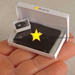 May include: Miniature black laptop with a yellow star logo, a matching mouse, and a white box labeled "cBook Pro." The laptop and mouse are inside a white box with a foam insert. The laptop is black and has a yellow star logo.