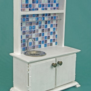 May include: A white miniature kitchen cabinet unit featuring a backsplash of blue, white, and purple square tiles. The unit includes a silver-toned sink with a faucet, a horizontal metal rod, and two cabinet doors with small round knobs.