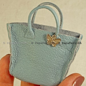 Pode incluir: Uma pequena bolsa de mão de couro azul claro com um pingente de borboleta dourada. A pequena bolsa tote tem duas alças arredondadas e uma superfície texturizada. O interior é de uma cor cinza contrastante.