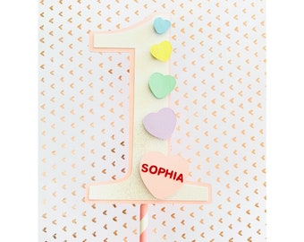 May include: A number one cake topper with pastel hearts and the name "SOPHIA" in red. The number is white with a pink outline and glitter accents. The topper is on a pink and white striped stick.