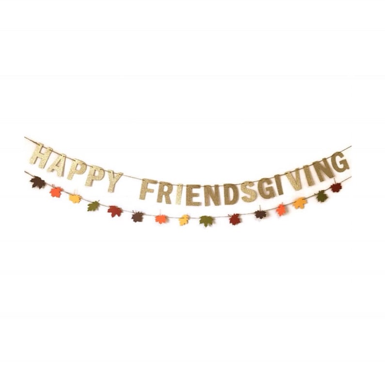 Może przedstawiać: Złota girlanda z błyszczącymi literami z napisem "Happy Friendsgiving" przewleczona na sznurku z małymi papierowymi liśćmi w odcieniach pomarańczowego, ż&oacute;łtego, czerwonego i brązowego.
