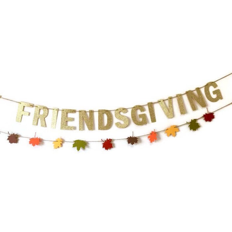 Może przedstawiać: Złota błyszcząca girlanda z napisem "FRIENDSGIVING" przewleczona na sznurku z małymi papierowymi liśćmi w odcieniach pomarańczowego, czerwonego i zielonego.