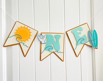 May include: A white and gold banner with the word "ONE" in blue and green letters. The banner has a sun, waves, and a hand making the shaka sign on it. The banner is perfect for a first birthday party.