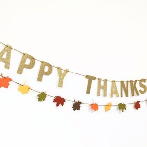 Może przedstawiać: Złota girlanda z błyszczącymi literami z napisem "Happy Thanksgiving" przewleczona przez brązowy sznurek i ozdobiona papierowymi liśćmi w jesiennych kolorach.