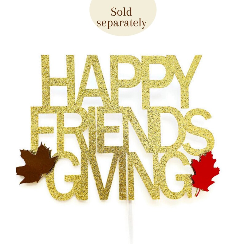 Może przedstawiać: Złoty topper na ciasto z błyszczącymi literami z napisem "Happy Friendsgiving" i dwoma jesiennymi liśćmi, jednym brązowym i jednym czerwonym.