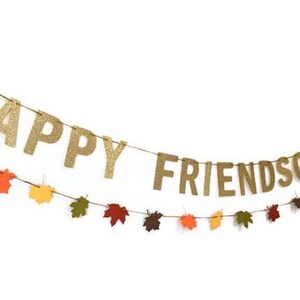 Może przedstawiać: Złota girlanda z błyszczącymi literami z napisem "Happy Friendsgiving" i ozdobami w kształcie jesiennych liści.