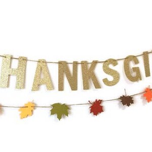 Może przedstawiać: Złota błyszcząca girlanda z okazji Dnia Dziękczynienia z napisem "THANKSGIVING" dużymi literami. Pod girlandą przewieszona jest wstążka z jesiennymi liśćmi w kolorze pomarańczowym, czerwonym, ż&oacute;łtym i brązowym.