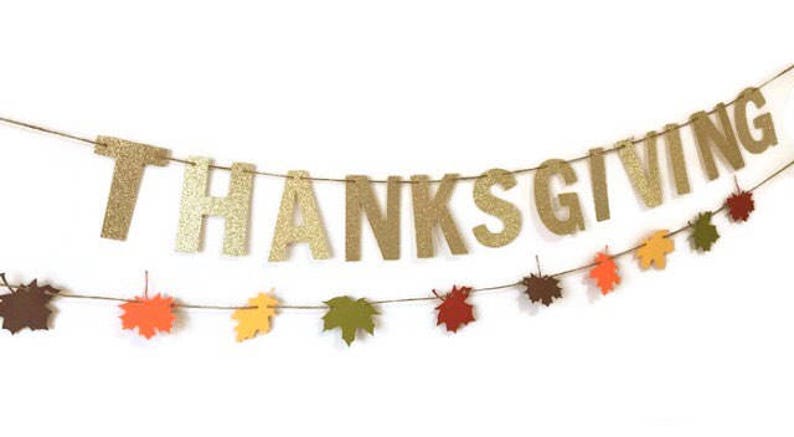 Może przedstawiać: Złota błyszcząca girlanda z okazji Dnia Dziękczynienia z napisem "THANKSGIVING" dużymi literami. Pod girlandą przewieszona jest wstążka z jesiennymi liśćmi w kolorze pomarańczowym, czerwonym, ż&oacute;łtym i brązowym.