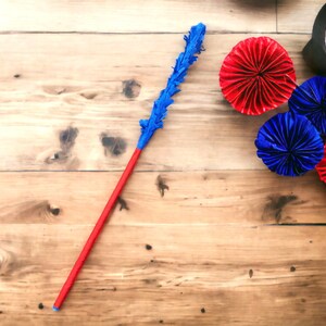 May include: A handmade red and blue decorative wand with a blue feathery top. The wand is lying on a wooden surface next to red and blue paper fans. The wand is a craft item.