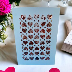 May include: A light blue card with a cut-out design of hearts filled with red glitter. The card is on a white surface with two pink hearts.