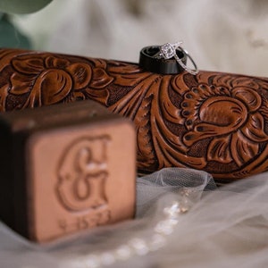 May include: Two wedding bands, one with a diamond, are placed on a brown leather box with a floral design. The box is sitting on a white fabric with a date engraved on it.