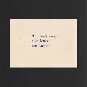 Op de afbeelding: Een beige kaart met de tekst 'Hij heeft voor elke letter een lachje.' gedrukt in donkerblauwe inkt.